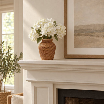 Woven rattan vase with a flared rim holding white hydrangeas on a white fireplace mantel beneath a framed landscape painting