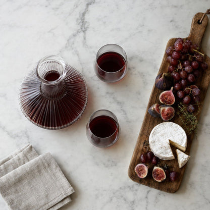 Overhead view of a round ribbed glass decanter with red wine and two glasses of red wine beside a wood board with cheese, grapes and figs on a white marble surface