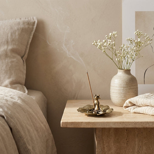 Decorative scene with a vase, incense burner, and flowers on a wooden table next to a bed.